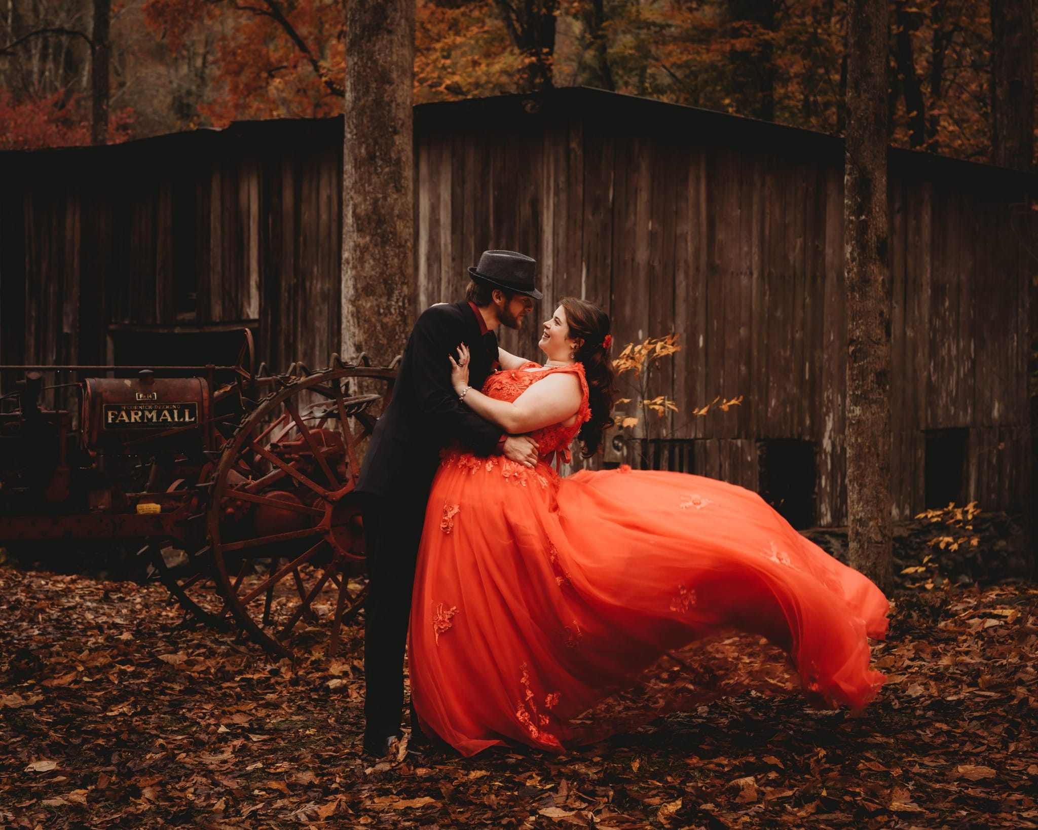 Couple embracing in black and a flowing orange gown beside a rustic wooden building.