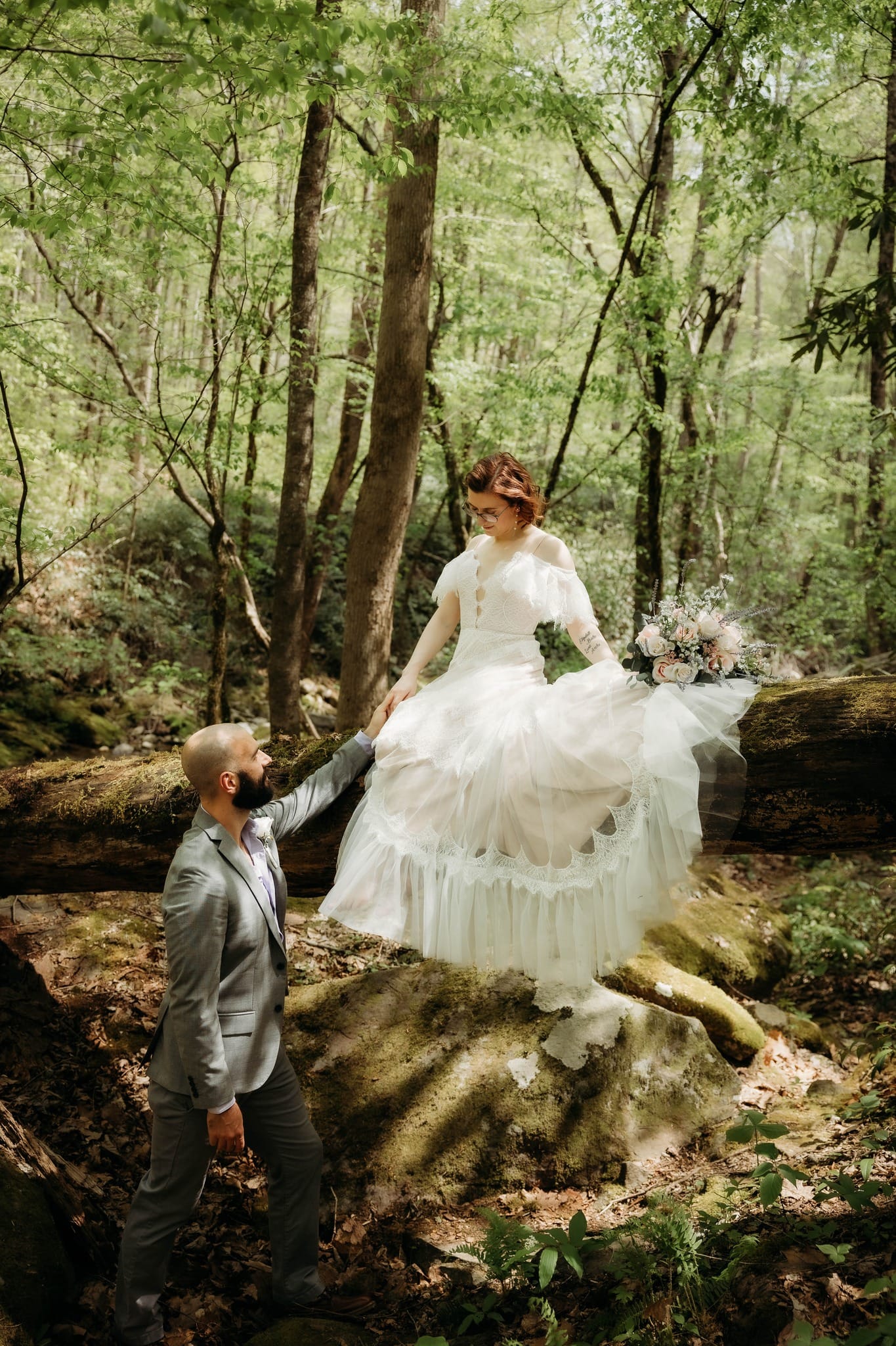 Bride in a ruffled white dress holding her partner’s hand beside a fallen tree.