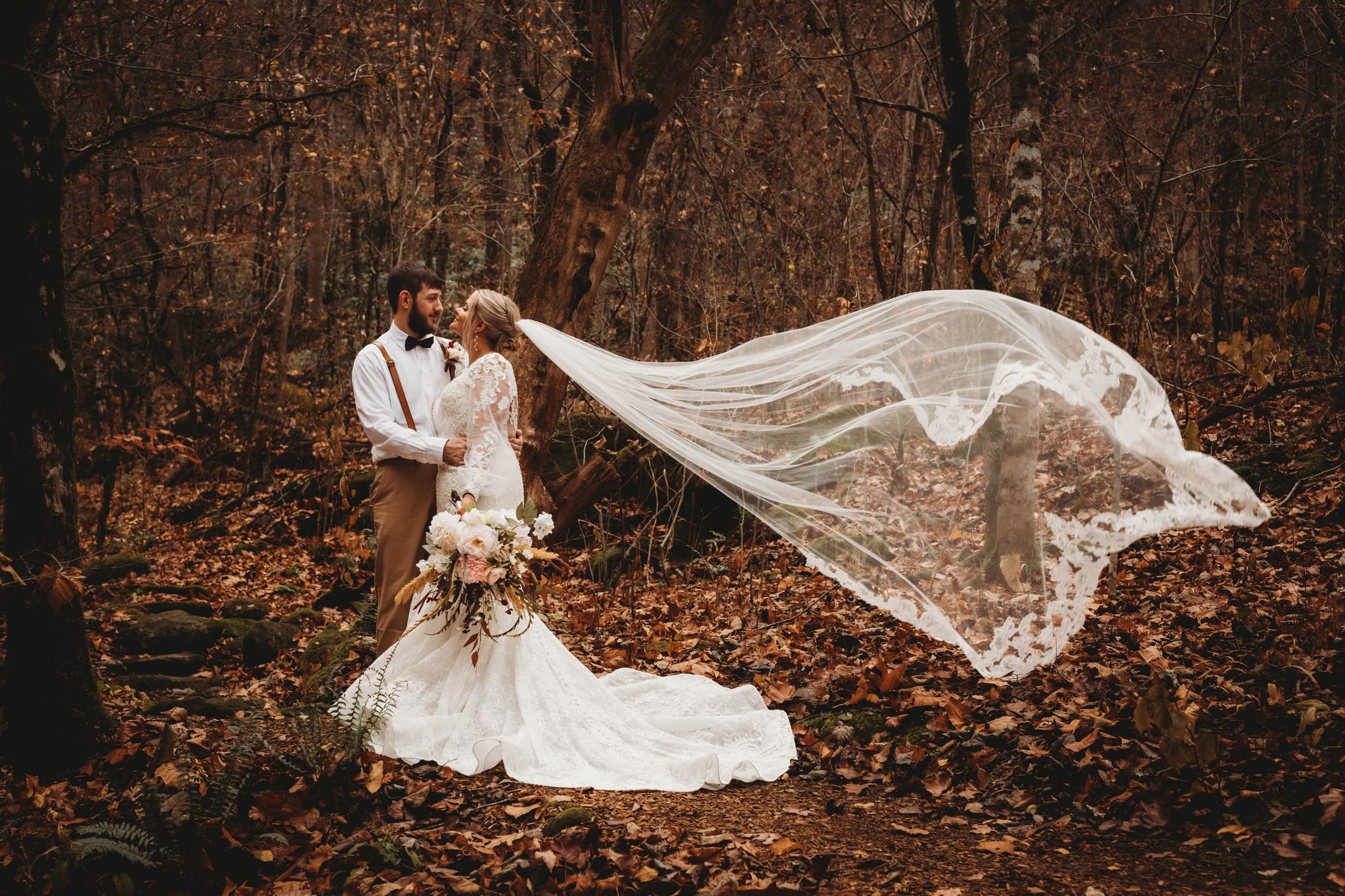 Wedding couple among autumn trees with a long lace veil floating behind them.
